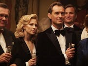 Blonde woman and man in formal attire stand holding drinks at a cocktail party.