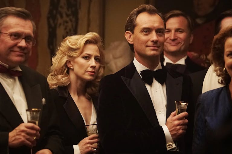 Blonde woman and man in formal attire stand holding drinks at a cocktail party. 