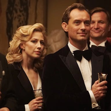 Blonde woman and man in formal attire stand holding drinks at a cocktail party. 