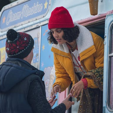 A young Black woman and young white man wearing thick coats and hats hold hands as the woman leans out the window of a frosty ice cream van