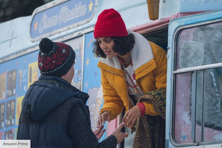 A young Black woman and young white man wearing thick coats and hats hold hands as the woman leans out the window of a frosty ice cream van