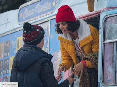 A young Black woman and young white man wearing thick coats and hats hold hands as the woman leans out the window of a frosty ice cream van