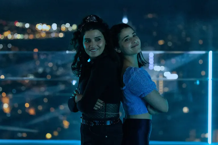 Two young women pose against a night skyline, with their arms folded standing back to back and smiling