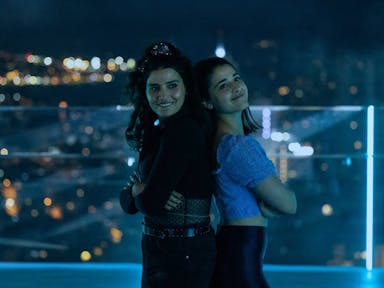Two young women pose against a night skyline, with their arms folded standing back to back and smiling
