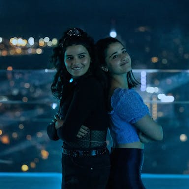 Two young women pose against a night skyline, with their arms folded standing back to back and smiling