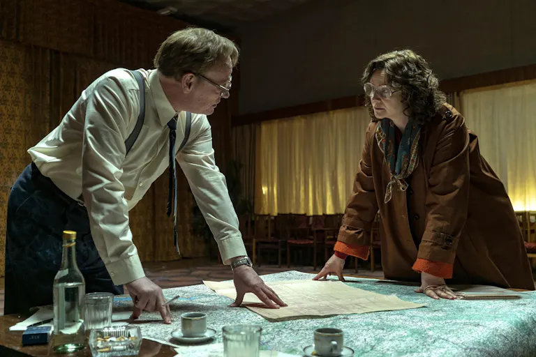 A white man and woman with thick lens glasses lean over a table with maps and documents, looking intense