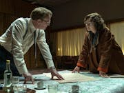 A white man and woman with thick lens glasses lean over a table with maps and documents, looking intense