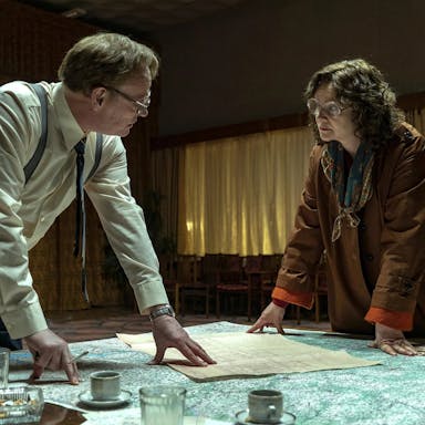 A white man and woman with thick lens glasses lean over a table with maps and documents, looking intense
