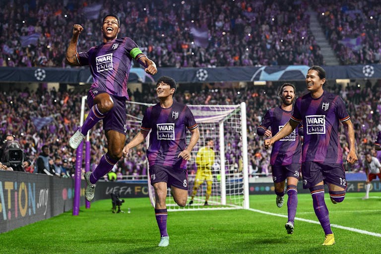 Game still of realistic male football players in purple kit running on the pitch celebrating with big crowds in the background