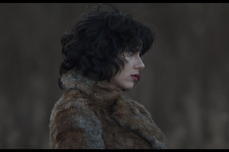 A pale woman with red lips and short wavy black hair in a dark fur coat, with a serious face, looks off into the distance