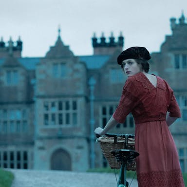 Young woman in old fashioned clothing, a black hat and red dress, stands with her bicycle in front of a huge stately home