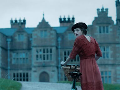Young woman in old fashioned clothing, a black hat and red dress, stands with her bicycle in front of a huge stately home