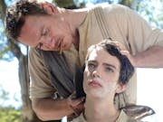 Man wearing a beige top with a cigar in his mouth cuts the hair of a teenage boy using a large old-fashioned knife.