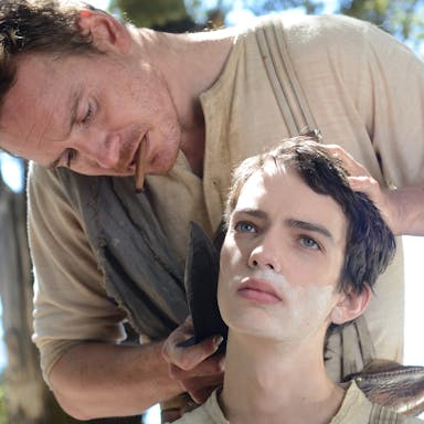 Man wearing a beige top with a cigar in his mouth cuts the hair of a teenage boy using a large old-fashioned knife. 