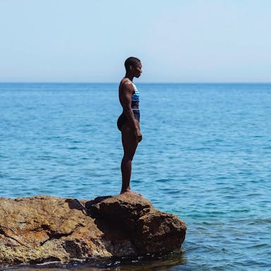 A black woman with cropped hair stands in a swim suit on the edge of a low rock overlooking a serene, vast blue sea in Greece