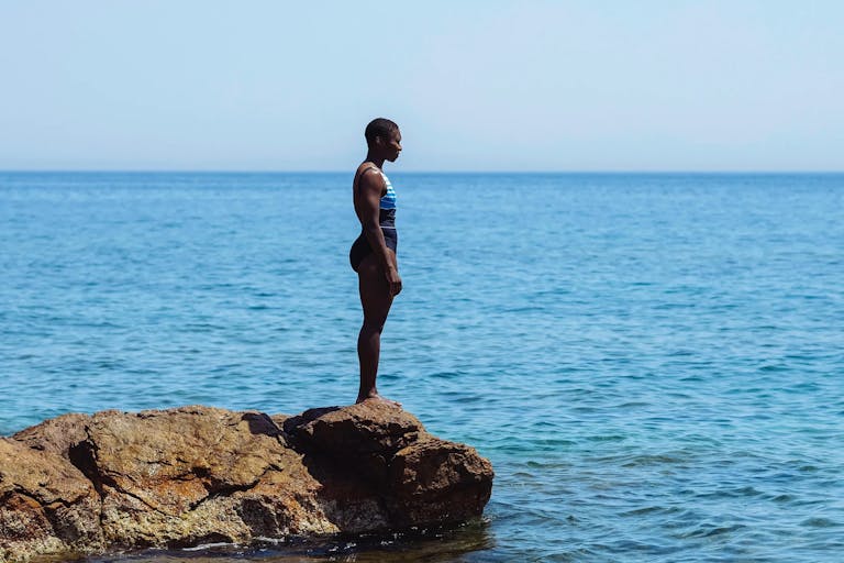 A black woman with cropped hair stands in a swim suit on the edge of a low rock overlooking a serene, vast blue sea in Greece