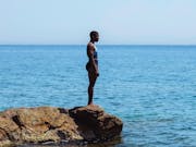 A black woman with cropped hair stands in a swim suit on the edge of a low rock overlooking a serene, vast blue sea in Greece