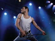 A lean man with dark hair and thick moustache, performing on stage in a power stance, wearing a white vest and jeans