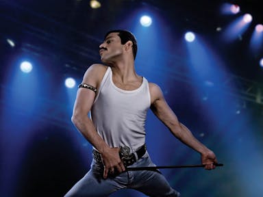A lean man with dark hair and thick moustache, performing on stage in a power stance, wearing a white vest and jeans 