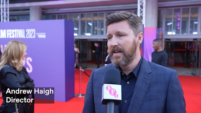 A white man with dark hair and beard speaks into a microphone on a red carpet