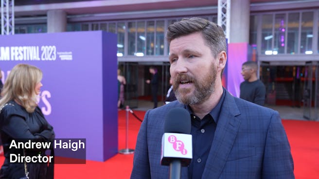 A white man with dark hair and beard speaks into a microphone on a red carpet
