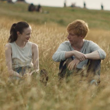 A white woman and a white man with ginger hair sit in a golden lit field of long brown grass smiling at each other