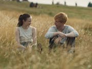 A white woman and a white man with ginger hair sit in a golden lit field of long brown grass smiling at each other