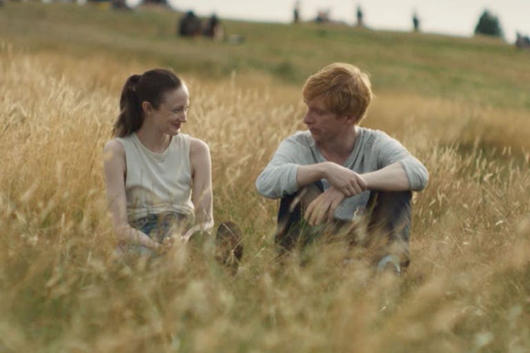 A white woman and a white man with ginger hair sit in a golden lit field of long brown grass smiling at each other