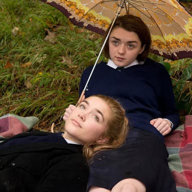 Two girls in school uniform recline on a picnic blanket on the grass. One of them holds an umbrella.