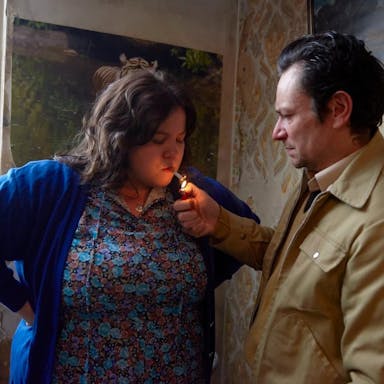 A man in a beige jacket lights a cigarette in a woman's mouth for her. 