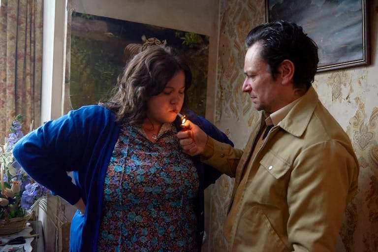 A man in a beige jacket lights a cigarette in a woman's mouth for her.