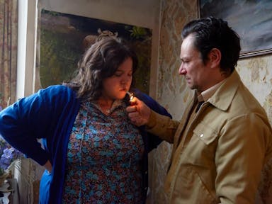 A man in a beige jacket lights a cigarette in a woman's mouth for her. 