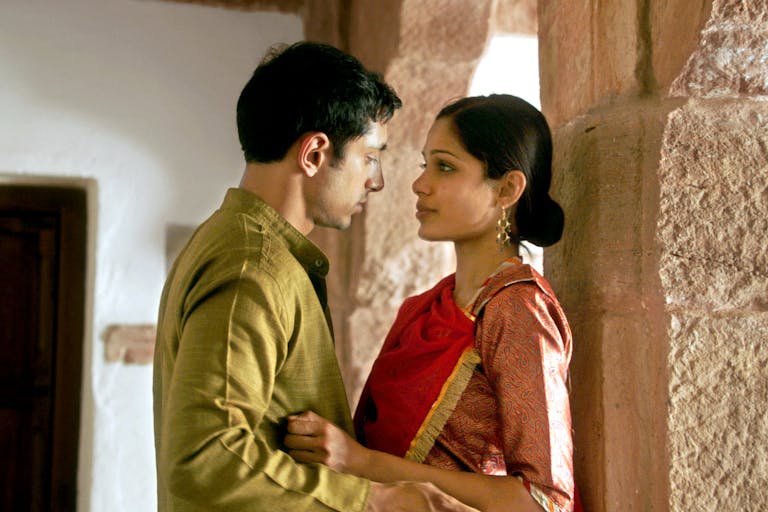 A dark haired man and woman in traditional Indian clothing leant against a stone pillar facing each other in an embrace