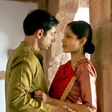 A dark haired man and woman in traditional Indian clothing leant against a stone pillar facing each other in an embrace