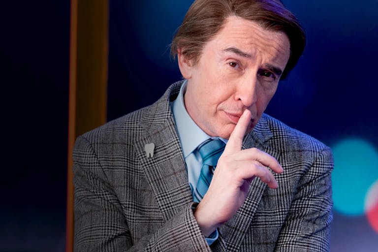 A middle aged white man in a suit pulling a smug face at the camera with a posed hand up to his mouth and eyebrow arched