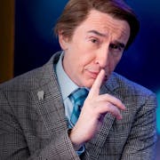 A middle aged white man in a suit pulling a smug face at the camera with a posed hand up to his mouth and eyebrow arched