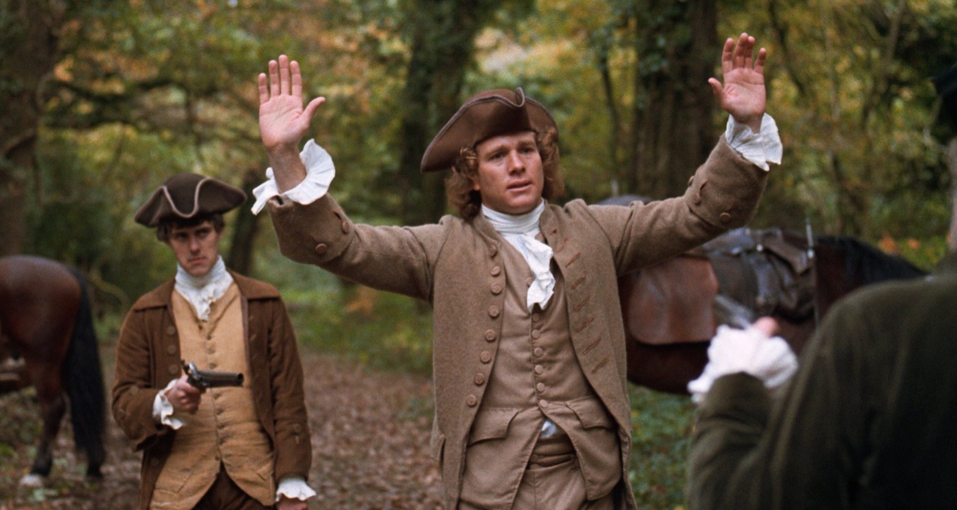 A white man in 18th Century attire lifting his hands up in a forest, whilst a weapon is being pointed at him from behind