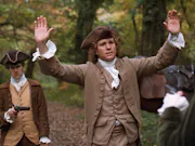 A white man in 18th Century attire lifting his hands up in a forest, whilst a weapon is being pointed at him from behind
