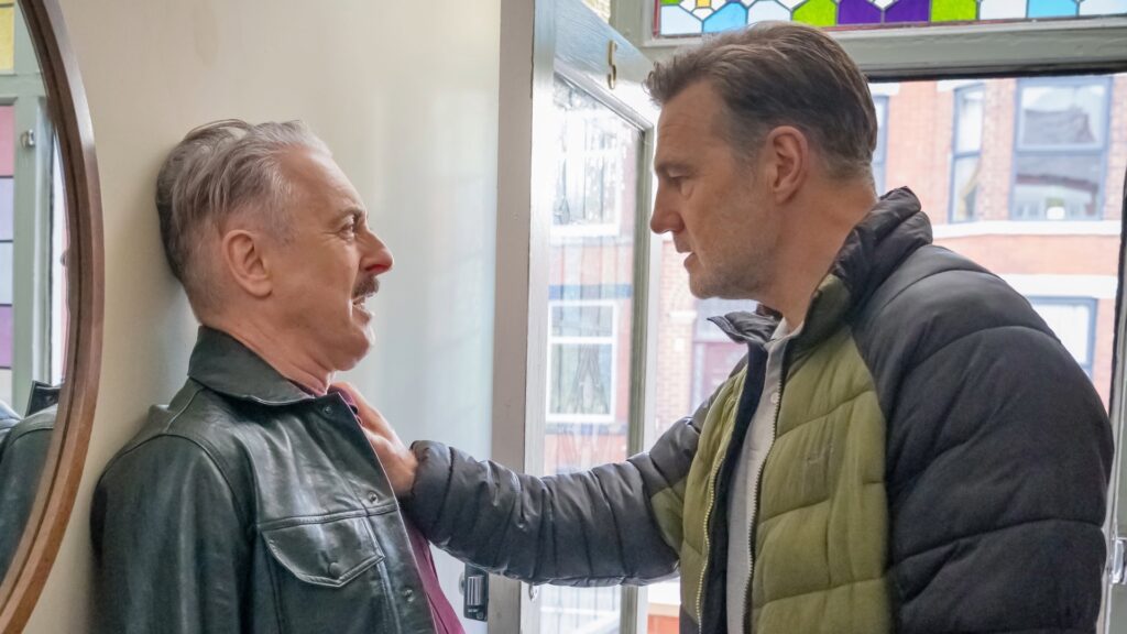 A tough-faced older white man pinning a scared older white man against a wall