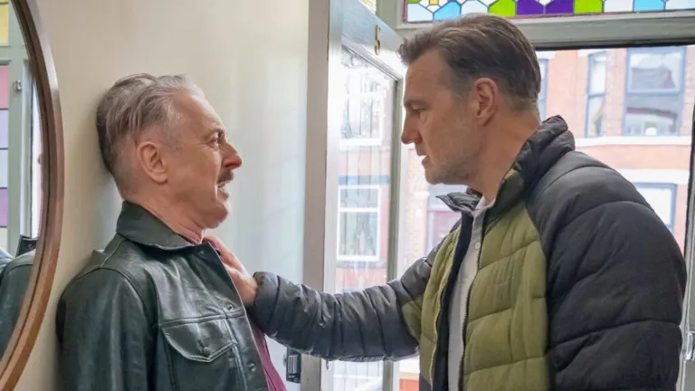 A tough-faced older white man pinning a scared older white man against a wall