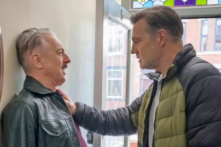 A tough-faced older white man pinning a scared older white man against a wall