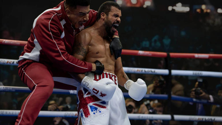 A shirtless and gloved black boxer mid-shout, with his coach on his back, also mid-shout