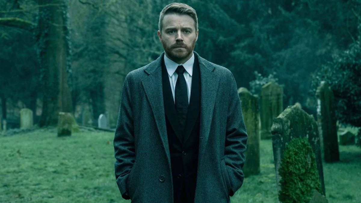 A middle-aged white man in a suit and overcoat, walking in a cemetery with a troubled look on his face