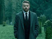 A middle-aged white man in a suit and overcoat, walking in a cemetery with a troubled look on his face