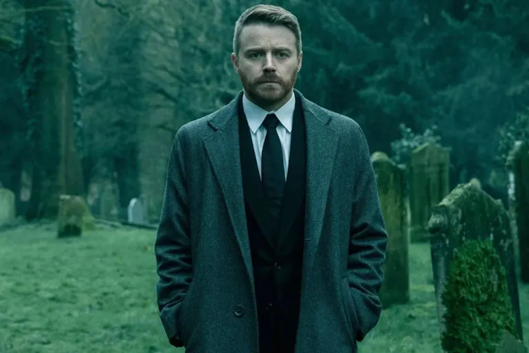 A middle-aged white man in a suit and overcoat, walking in a cemetery with a troubled look on his face