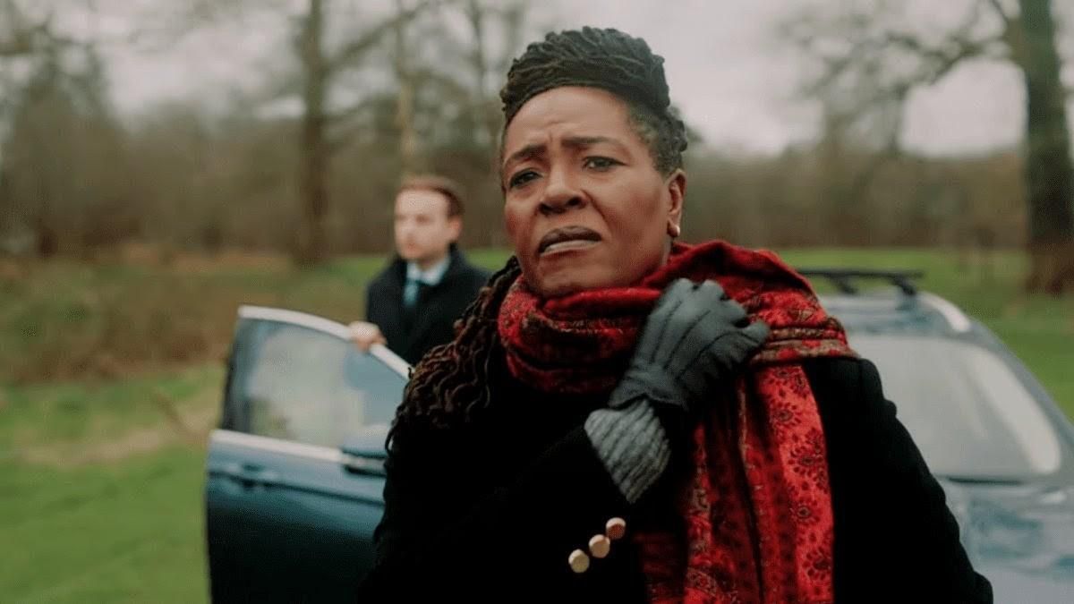 A black woman with black locs and a trench coat holding her scarf in place as she looks off into the distance. Behind her, a white man getting out of a car