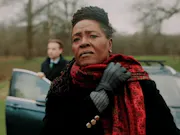 A black woman with black locs and a trench coat holding her scarf in place as she looks off into the distance. Behind her, a white man getting out of a car