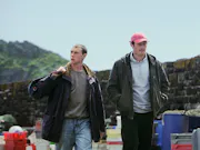 Two early 30's white men walking next to eachother away from hills in the background, both of which wearing a dark jacket. One looking down, the other (holding a bag) looking away