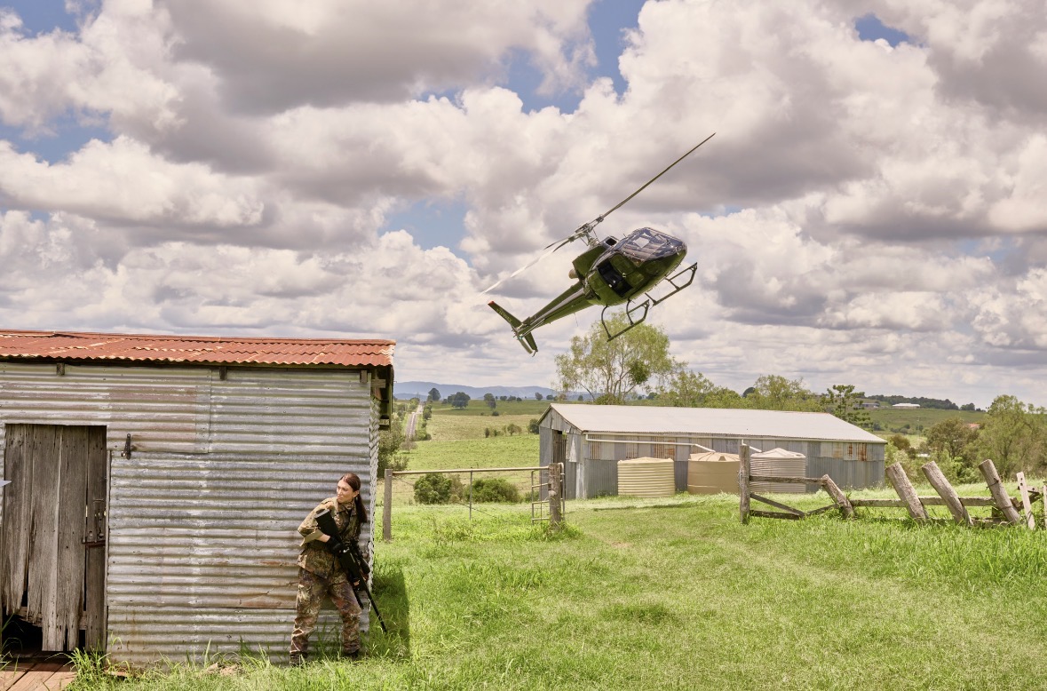 A woman wearing combat gear whilst hiding behind a shed in a field. In the background, a hellycopter in the sky