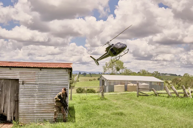 A woman wearing combat gear whilst hiding behind a shed in a field. In the background, a hellycopter in the sky